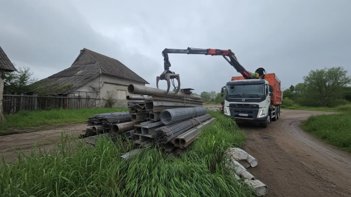 Кран-манипулятор грузит металлические трубы у деревенского дома