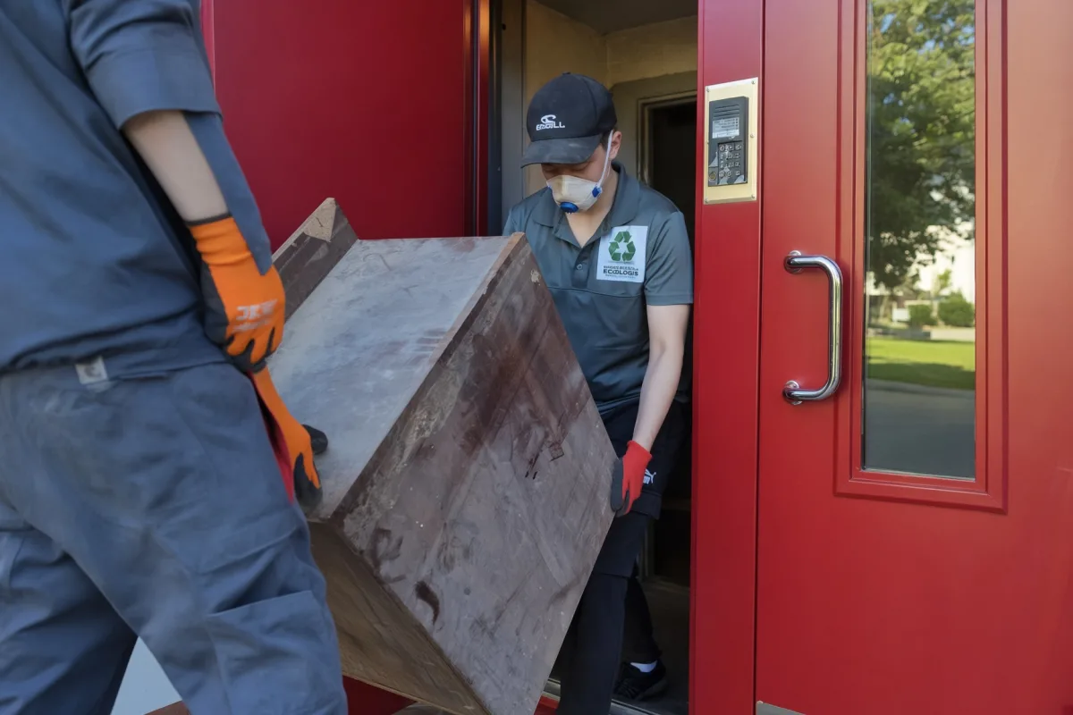 Грузчик в маске выносит деревянный комод из подъезда